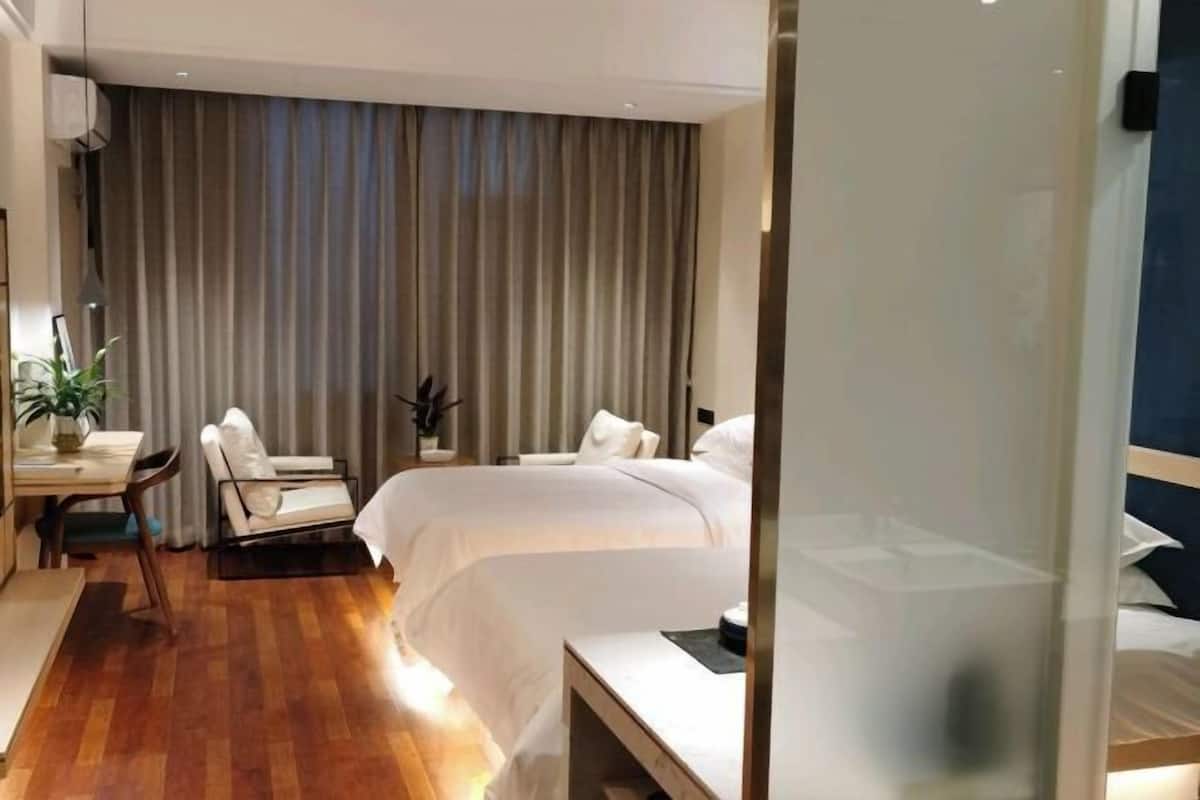 Una habitación de hotel moderna con piso de madera, una cama grande, un escritorio con silla y un ventanal con cortinas.