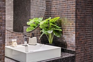 Une salle de bain avec un lavabo blanc, un miroir, une plante en pot et un mur carrelé.