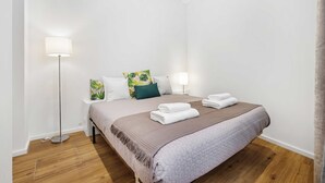 A neatly made bed with a brown quilt, a floor lamp, and a bedside table with a lamp. - Funchal Tropical - Filodendro by Homie (Funchal)