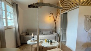 Une pièce chaleureuse avec un plancher en bois, un fauteuil suspendu, deux tables rondes ornées d’objets décoratifs, un canapé et une fenêtre avec des rideaux.