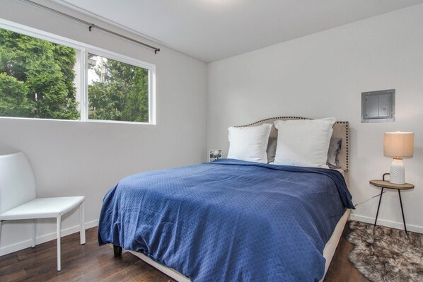 Una camera da letto con un letto, una sedia, una lampada e vista su alberi attraverso una finestra.