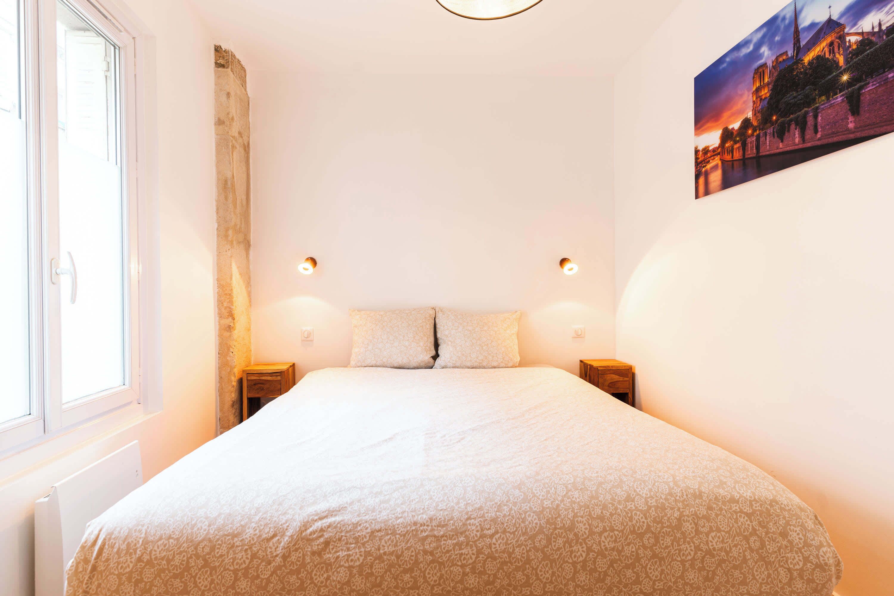 A bedroom with a large bed, two bedside tables, a window, and a wall-mounted picture.