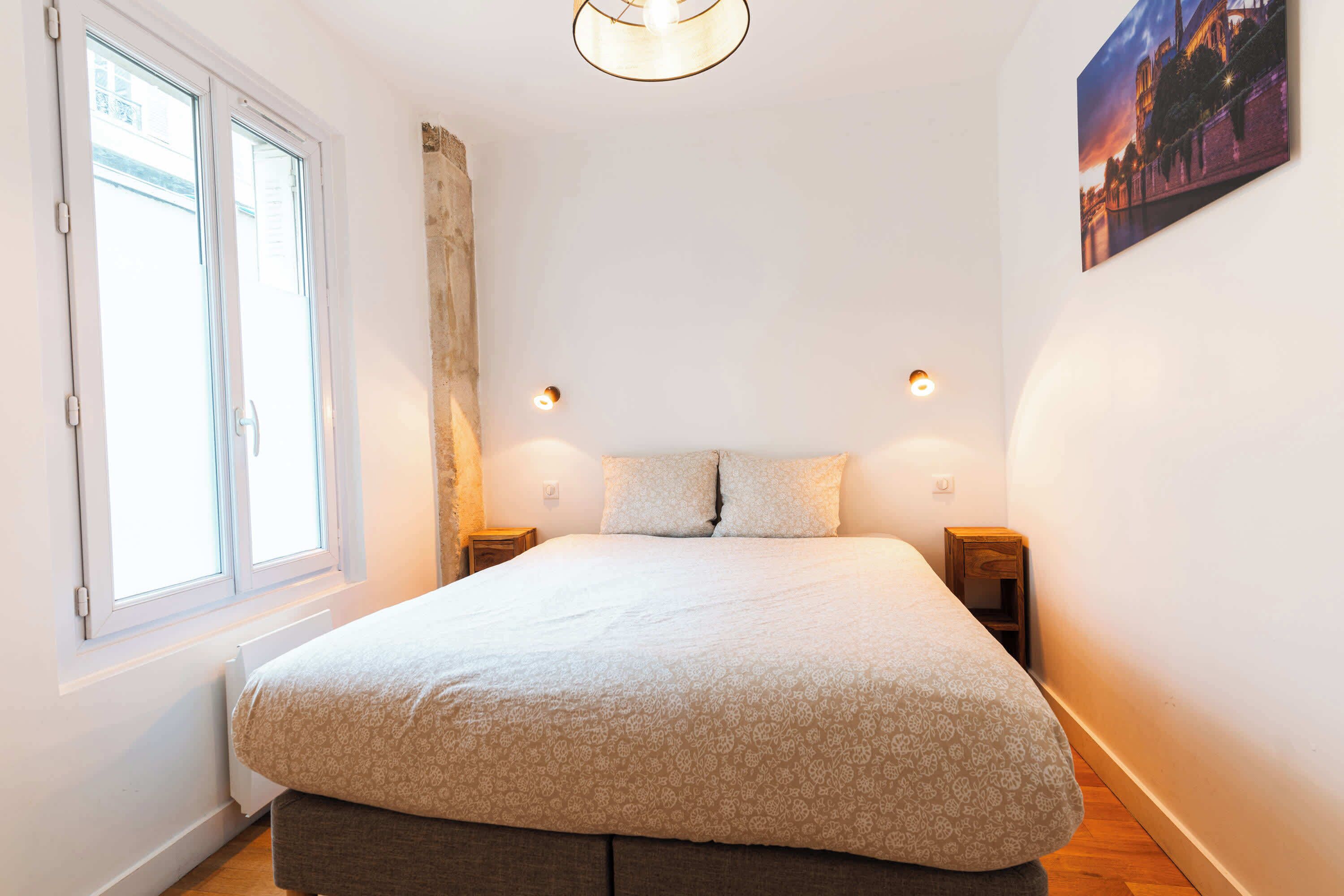 A bedroom with a large bed, a window with curtains, a night light, and a painting on the wall.