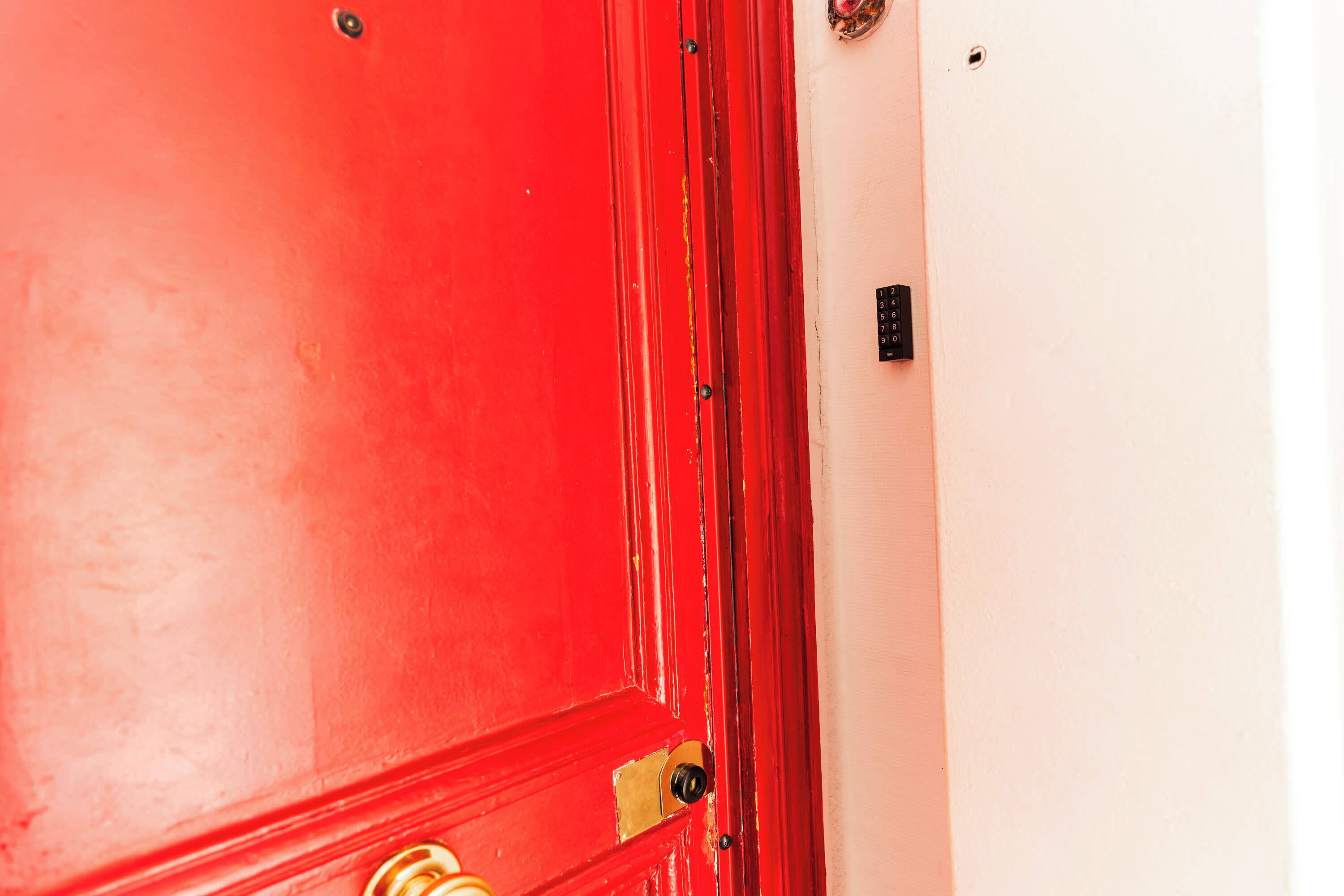 A red door with a brass doorknob and a keyhole, adjacent to a white wall with a black security panel.
