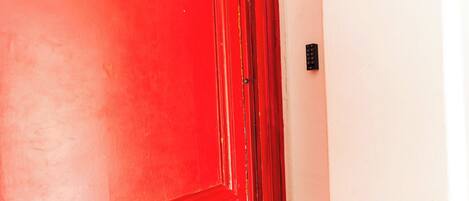 A red door with a brass doorknob and a keyhole, adjacent to a white wall with a black security panel.