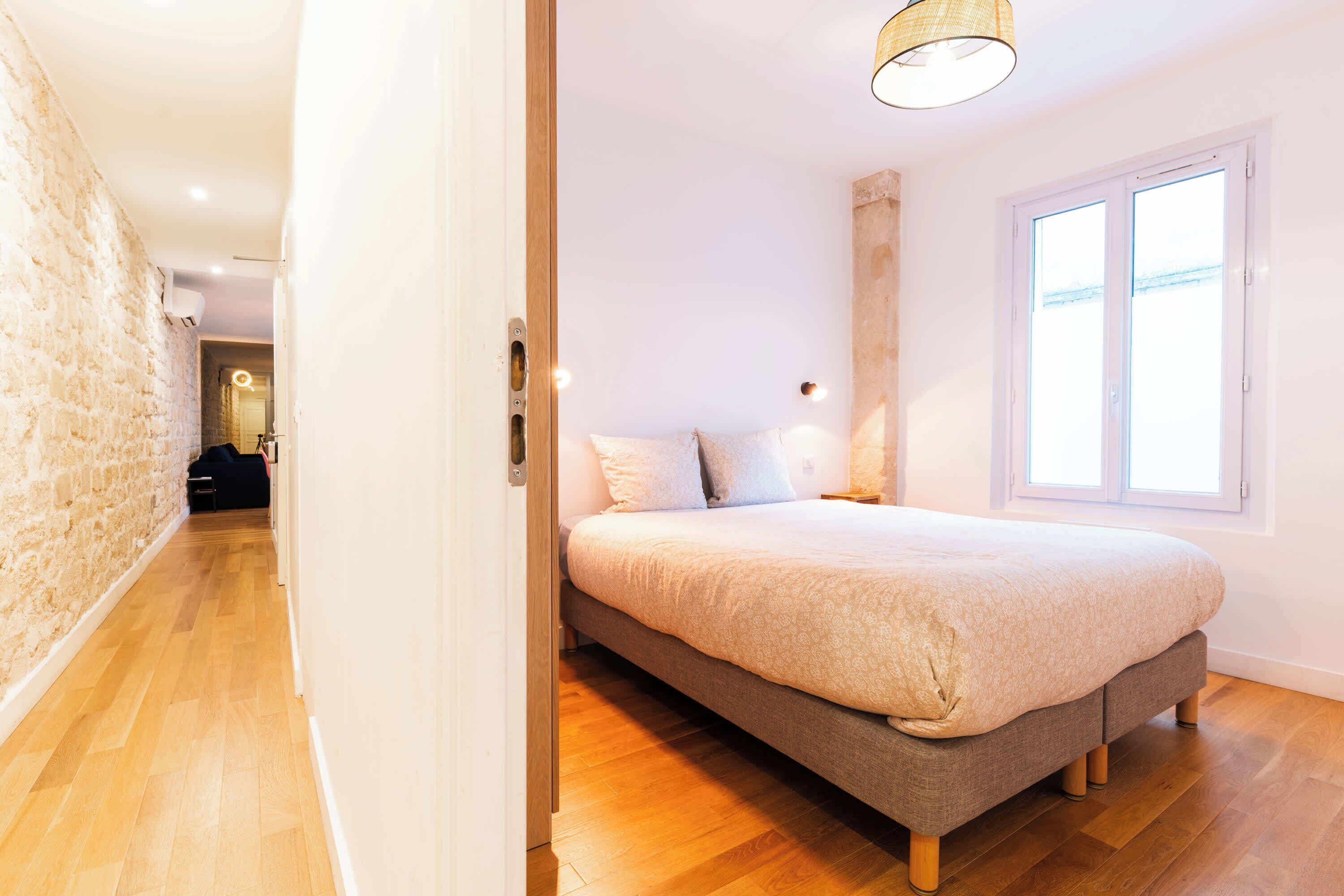 Une chambre à coucher comprenant un lit, un plancher en bois, une fenêtre et une porte donnant sur une autre pièce.