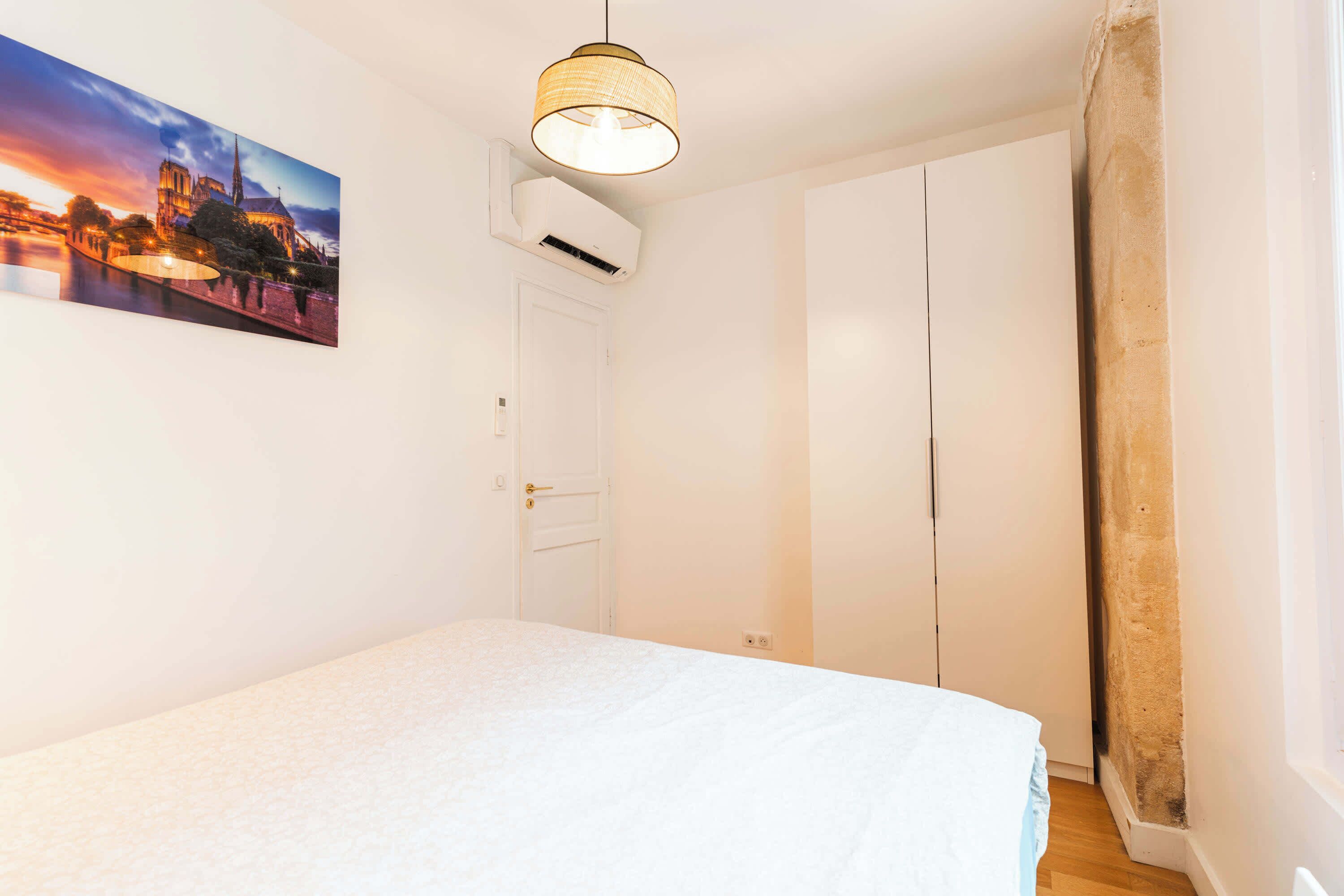 A bedroom with a bed, a painting of a cityscape, a ceiling light, and a white door.