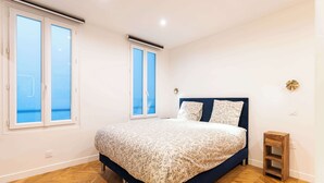 A bedroom with a large bed, two windows with blinds, a small wooden stool, and a wall-mounted light fixture. - Town House Nation, Bastille, AC by LCC Agency (Paris)