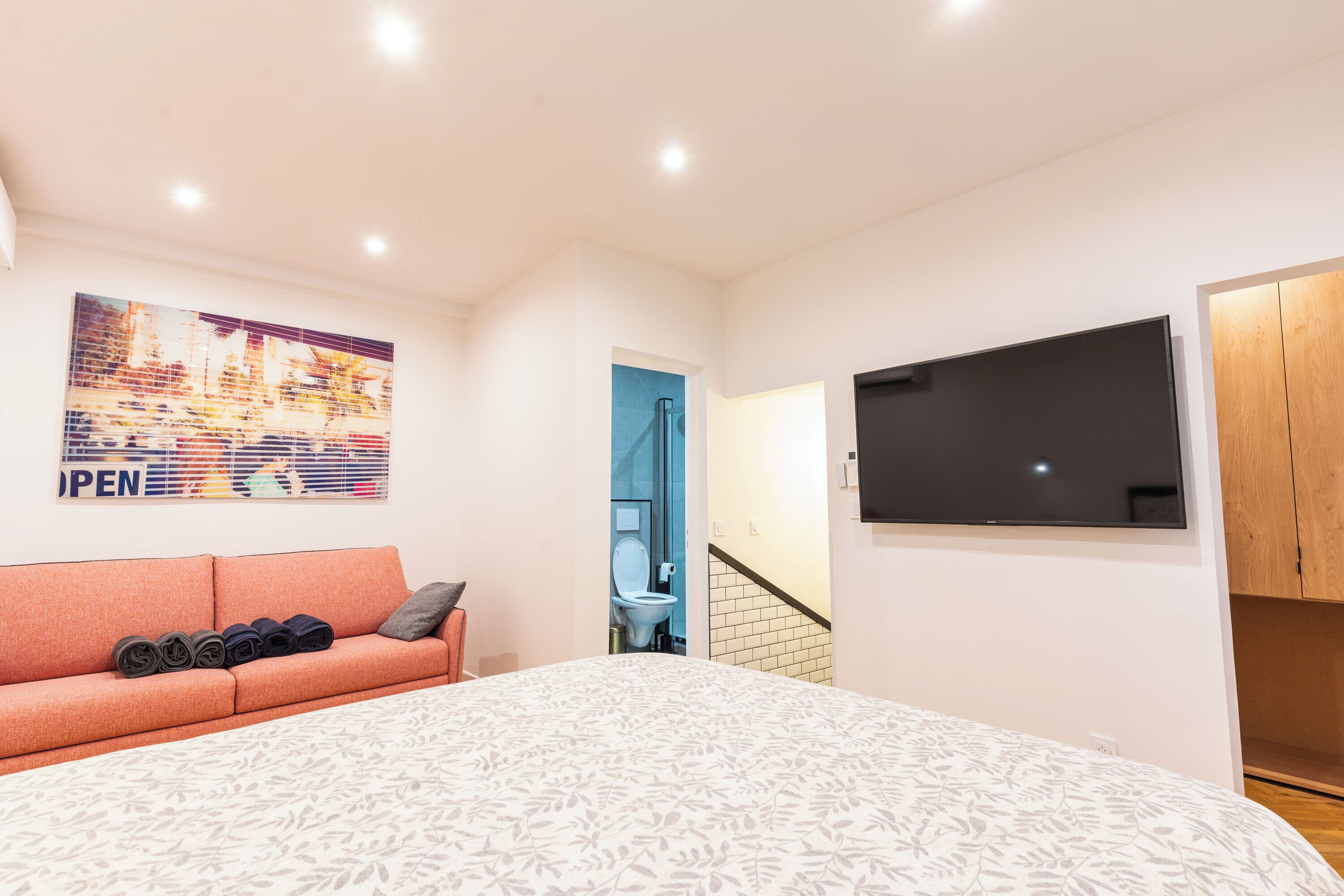 Une chambre avec un lit, un canapé sur lequel sont posés des objets pliés, un téléviseur accroché au mur et une salle de bain visible à travers une porte ouverte.
