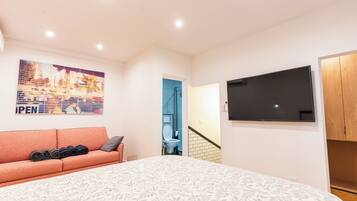 Une chambre avec un lit, un canapé sur lequel sont posés des objets pliés, un téléviseur accroché au mur et une salle de bain visible à travers une porte ouverte.