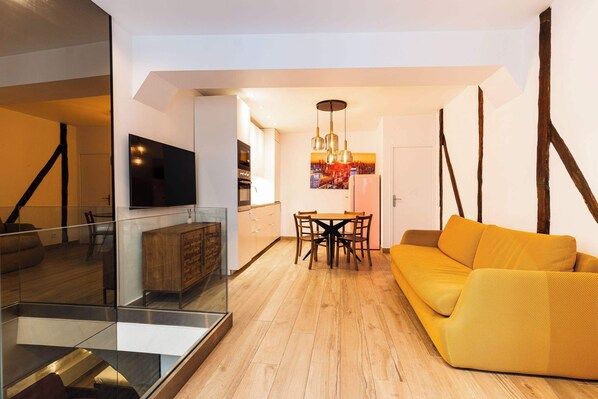 A modern living room with a wooden floor, a yellow sofa, a dining area with a table and chairs, and a kitchen in the background. - Loft 1 BR Marais, Notre Dame AC by LCC Agency (Paris)