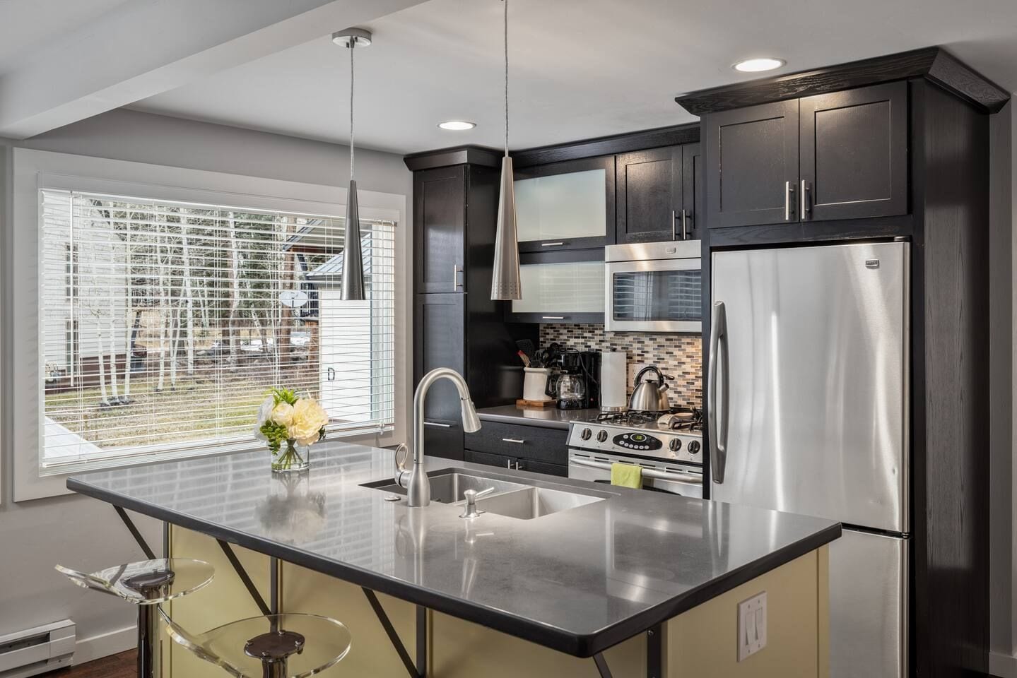 A modern kitchen with stainless steel appliances, a large window with blinds, and a bar area with stools.