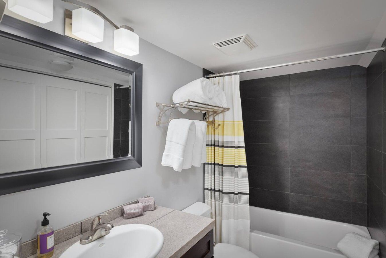 A modern bathroom with a large mirror, a bathtub, a shower with a striped curtain, and a vanity with a sink and towel dispensers.