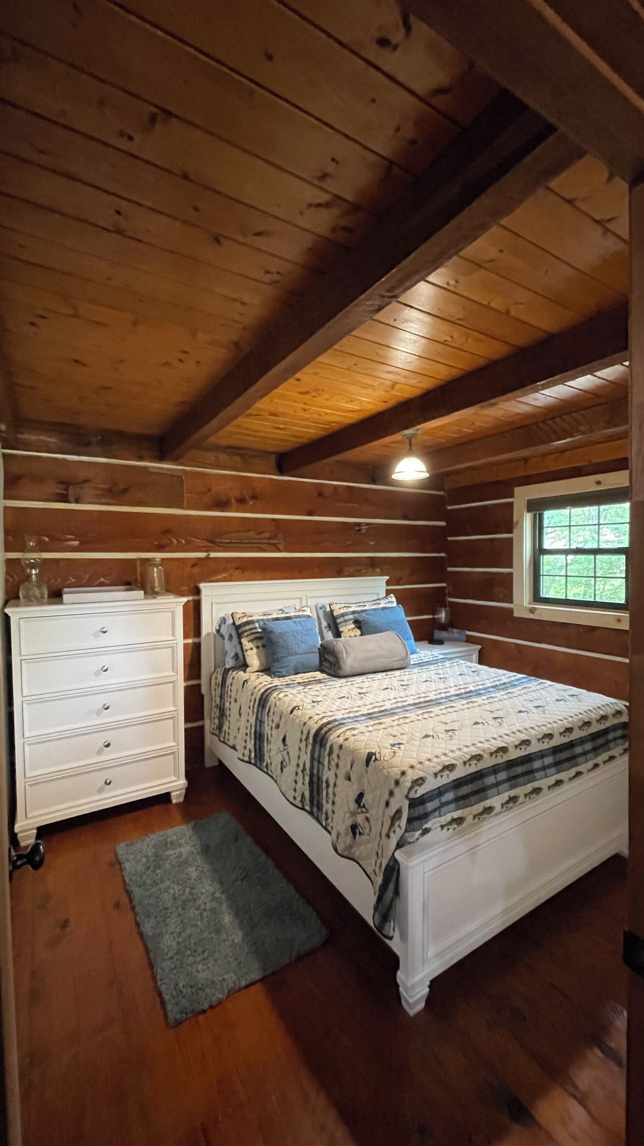 Un dormitorio con techo de madera, una cama con ropa de cama estampada, un aparador y una ventana con vistas a zonas verdes.