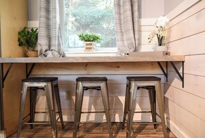 Une cuisine avec un plan de travail en bois, trois tabourets en métal, une fenêtre avec des rideaux et une petite plante en pot.