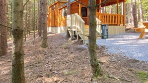 A cabin with a wooden deck and a covered porch, surrounded by tall trees in a forest.
