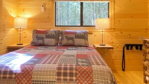 A cozy log cabin bedroom with a large bed, two bedside lamps, a window with a view of trees, and a ceiling fan.