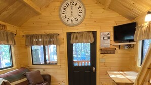 A cozy cabin interior with a wooden ceiling, a clock, a couch, a dining table, and a window with curtains.