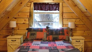 Ein gemütliches Schlafzimmer im Dachgeschoss mit schrägen Wänden, einem Bett mit Patchworkdecke, zwei Nachttischen mit Lampen und einem Fenster mit Karomustert-Vorhängen.