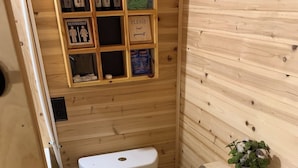 A small, wooden bathroom with a toilet, a shelf with toiletries, and a small plant. - The Hideaway near Cook Forest State Park (Leeper)