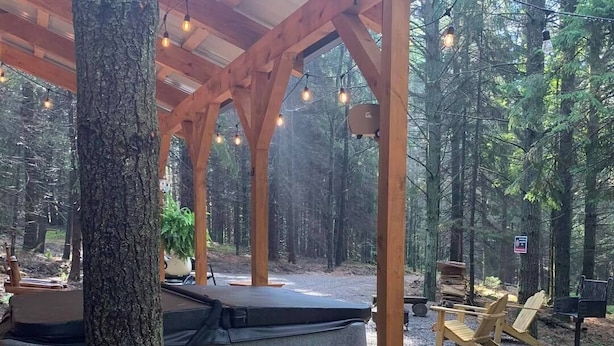 A covered outdoor seating area with a hot tub, wooden deck, and forest backdrop. - The Hideaway near Cook Forest State Park (Leeper)