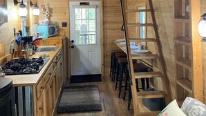 A compact kitchen with a stove, sink, and microwave. There is a door with a window, a ladder, and a shelf with dishes. - The Hideaway near Cook Forest State Park (Leeper)