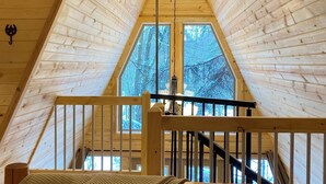 A wooden attic bedroom with a sloped ceiling, a ceiling fan, a window with a view of trees, and a wooden staircase. - The Treehouse near Cook Forest State Park (Leeper)