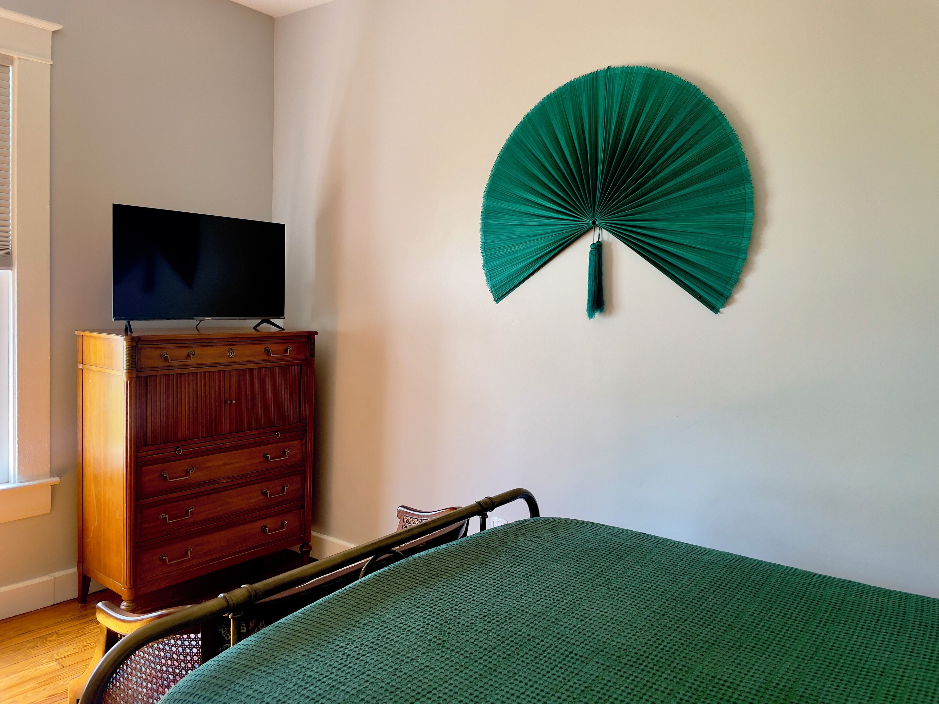 A bedroom with a bed, a wooden dresser, a television on top, and a green fan on the wall.
