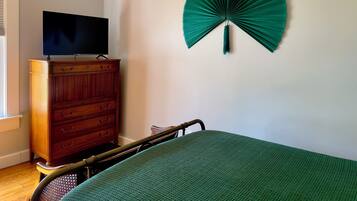 A bedroom with a bed, a wooden dresser, a television on top, and a green fan on the wall.