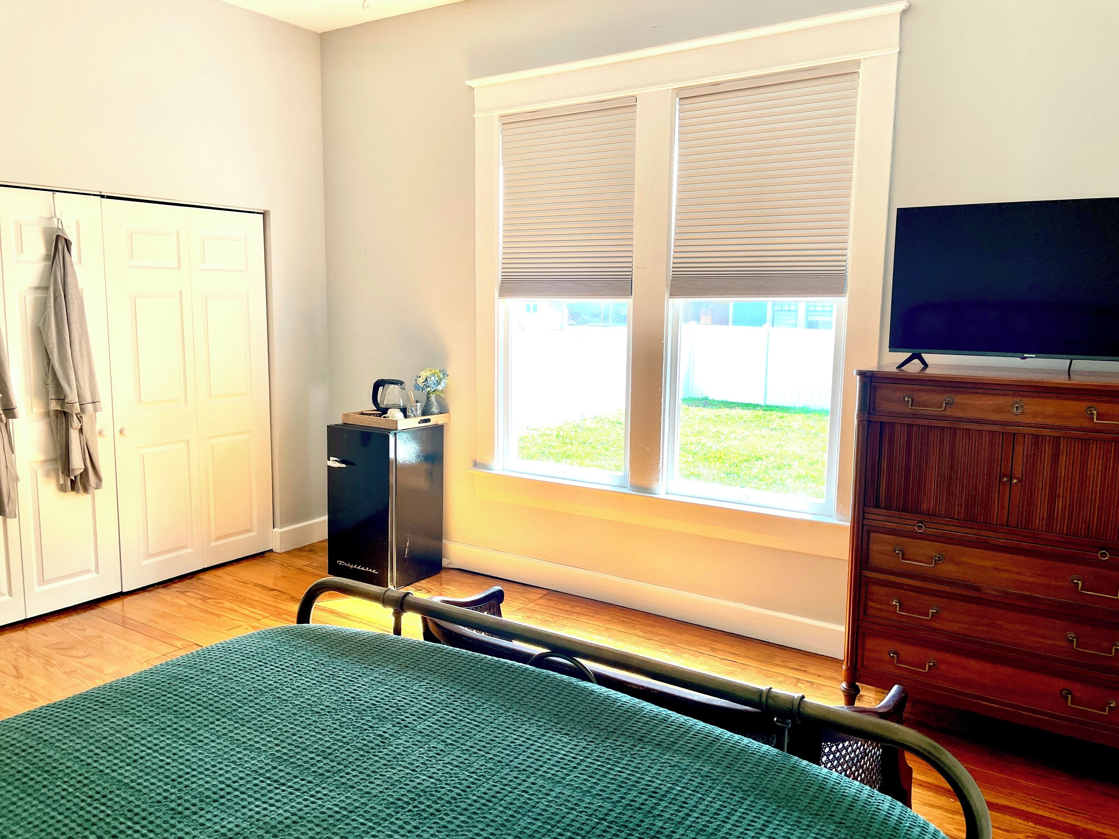 A bedroom with a bed, a dresser, a television, a window with blinds, and a door.
