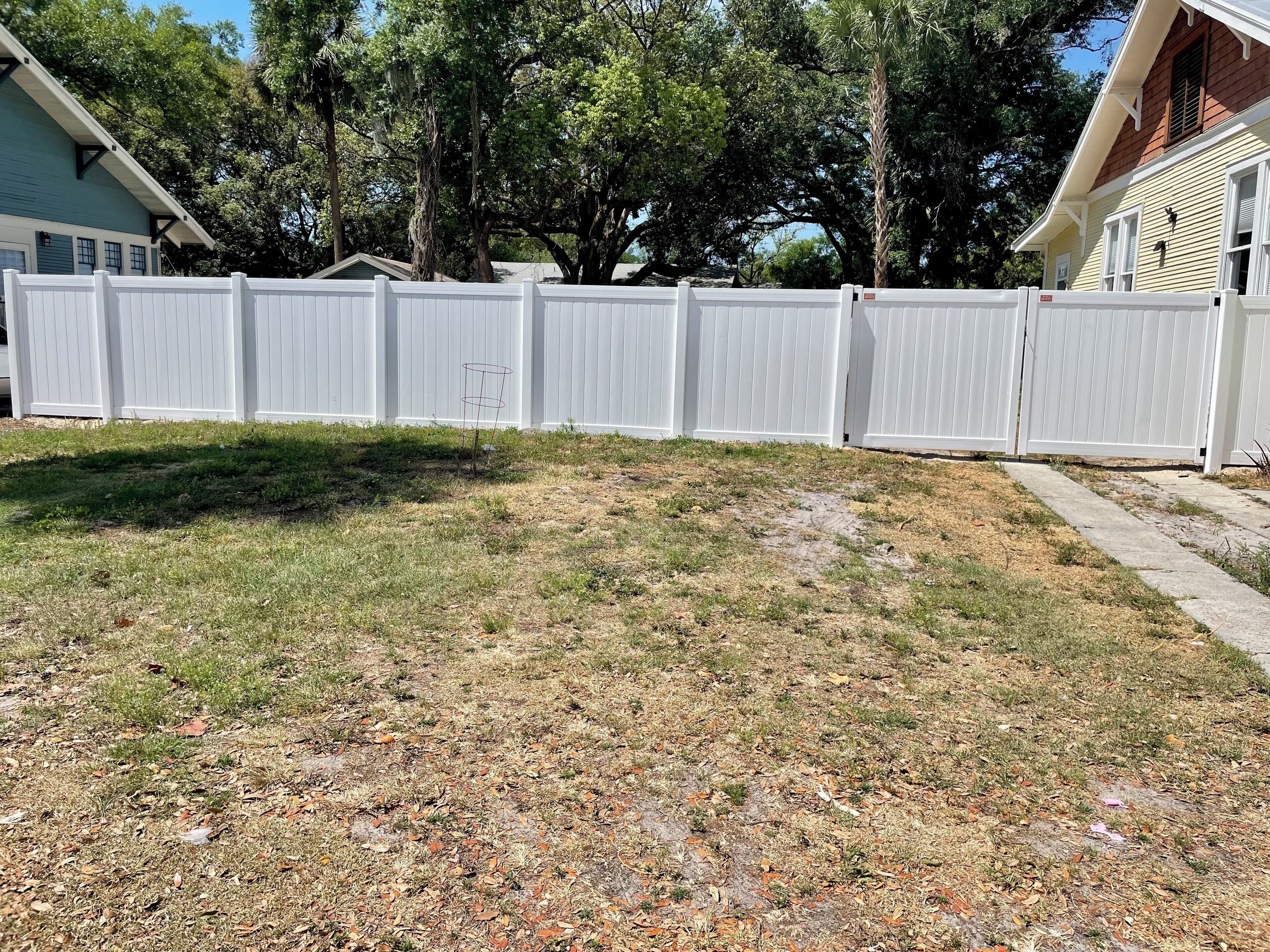 A fenced yard with a mix of grass and bare ground, surrounded by trees and houses.