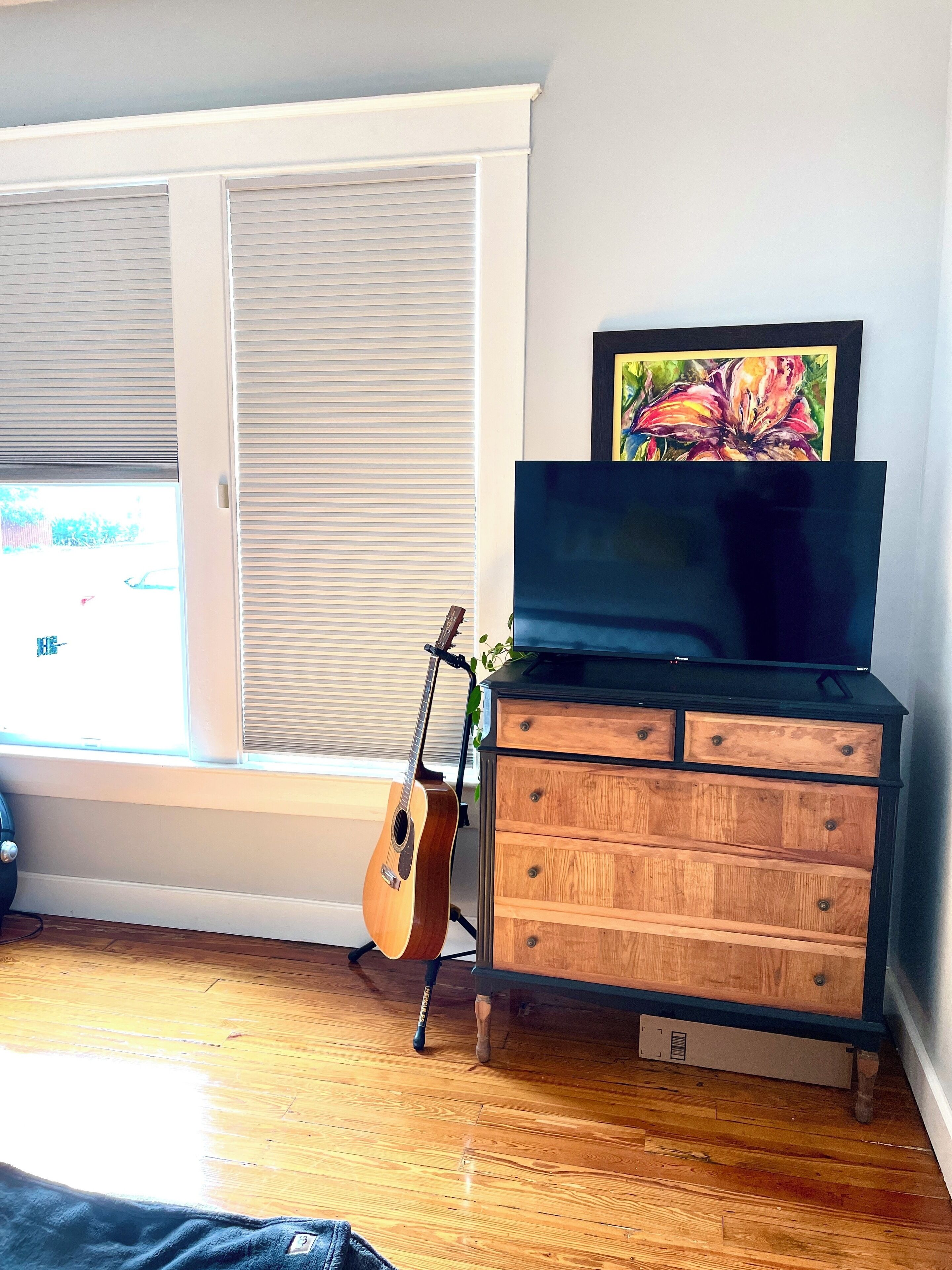 Une pièce avec une commode en bois, une guitare, un téléviseur posé sur le dessus, et une fenêtre avec des stores.