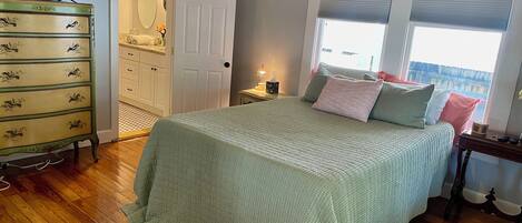 Un dormitorio con cama, piso de madera, cómoda, televisor y una puerta que da a un baño.