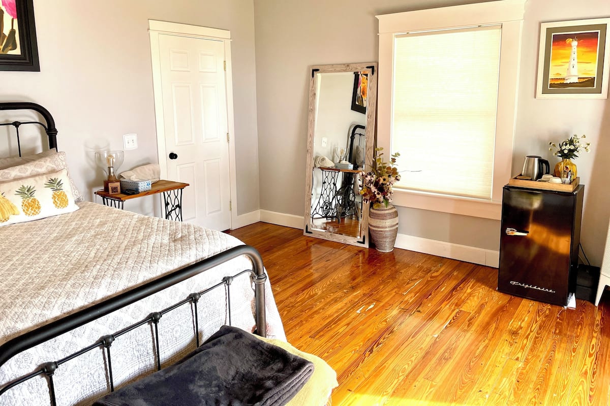 A bedroom with a bed, a dresser, a mirror, a window with blinds, and a small table with a lamp.