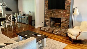 Una sala de estar con chimenea de ladrillo, un televisor montado encima y un comedor al fondo.