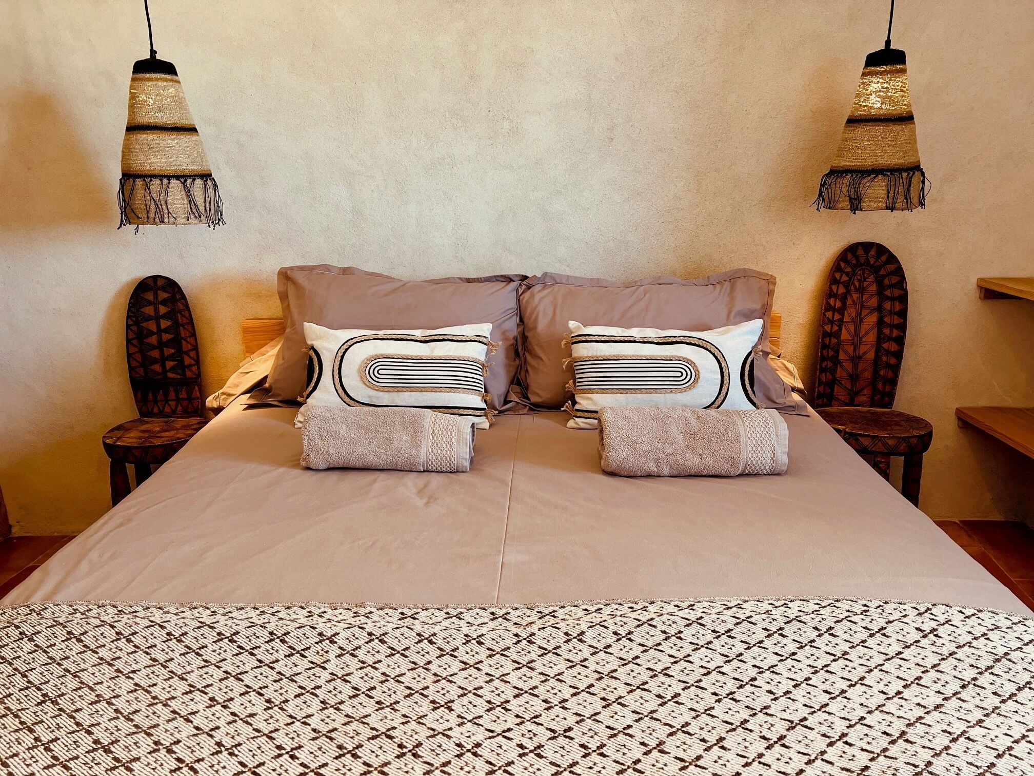 Cama bem arrumada com roupa de cama estampada, duas almofadas e um cobertor dobrado. Há duas mesas de cabeceira de madeira com abajures, e uma cadeira com encosto geométrico.