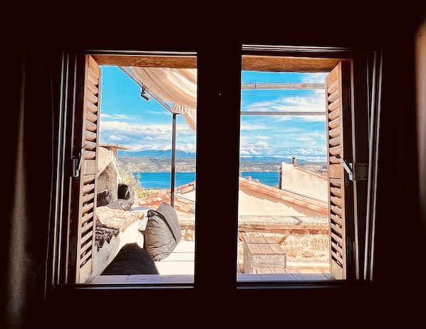 Vista a través de una ventana con contraventanas de madera, mostrando una terraza con pared de piedra y cielo azul despejado.