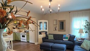 A living room with a dark blue sofa, a dining table with a vase of flowers, a kitchen with a white refrigerator and stove, and a chandelier.