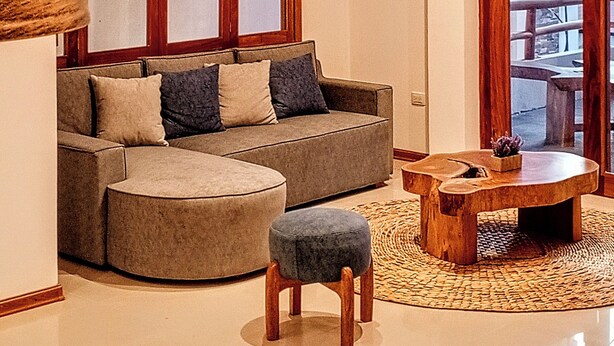 A modern living room with a sofa, ottoman, and coffee table, large windows, and a wooden ceiling beam. - Galapagos Coliving Ecuador Casa Tuna (Puerto Ayora)