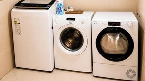 A laundry area with a compact washer and dryer, a small table with a bottle of detergent, and a wall-mounted control panel. - Buenos Aires de Ezeiza Resort Vacacional - Departamento N°1 (La Unión)