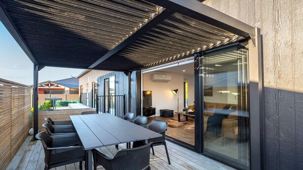 Een buitenterras met houten dek, een lange tafel met stoelen en een glazen deur naar een binnenruimte met een bank en een lamp.