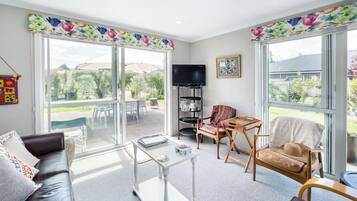 A spacious living room with a sofa, coffee table, and chairs, large windows with a view of the outdoors, and a television on a stand.