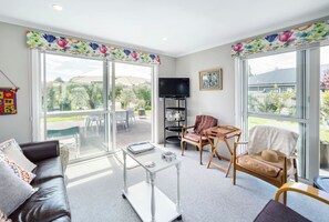 A spacious living room with a sofa, coffee table, and chairs, large windows with a view of the outdoors, and a television on a stand.