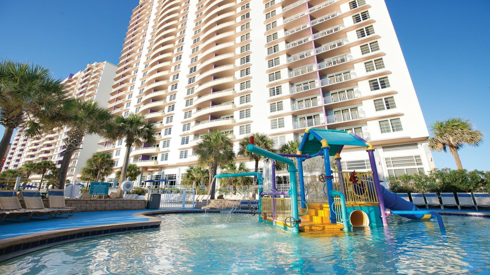 A hotel with a swimming pool, a children's play area, and palm trees.