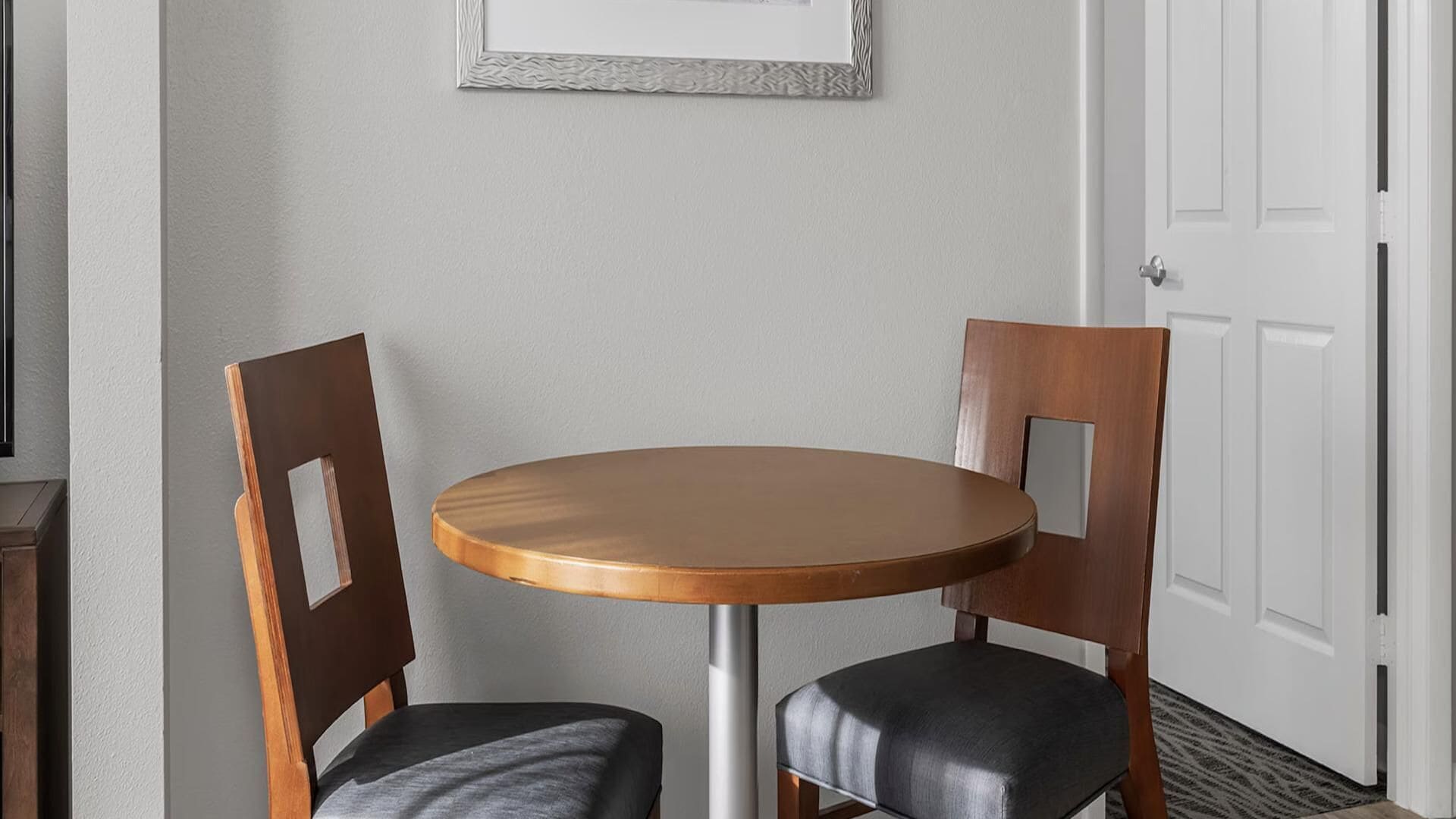 Una pequeña mesa redonda con dos sillas de madera, un cuadro enmarcado en la pared y una puerta blanca al fondo.