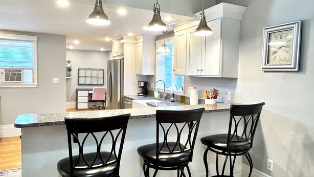 A modern kitchen with white cabinets, a stainless steel refrigerator, and a bar area with three black bar stools. - Stylish home next to train to Boston, near Salem (Wakefield)