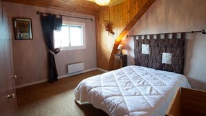 A bedroom with a wooden ceiling, a bed with a white quilt, a window with curtains, and a wall-mounted lamp. - Apartment 4/6 persons (AUMONT-AUBRAC)