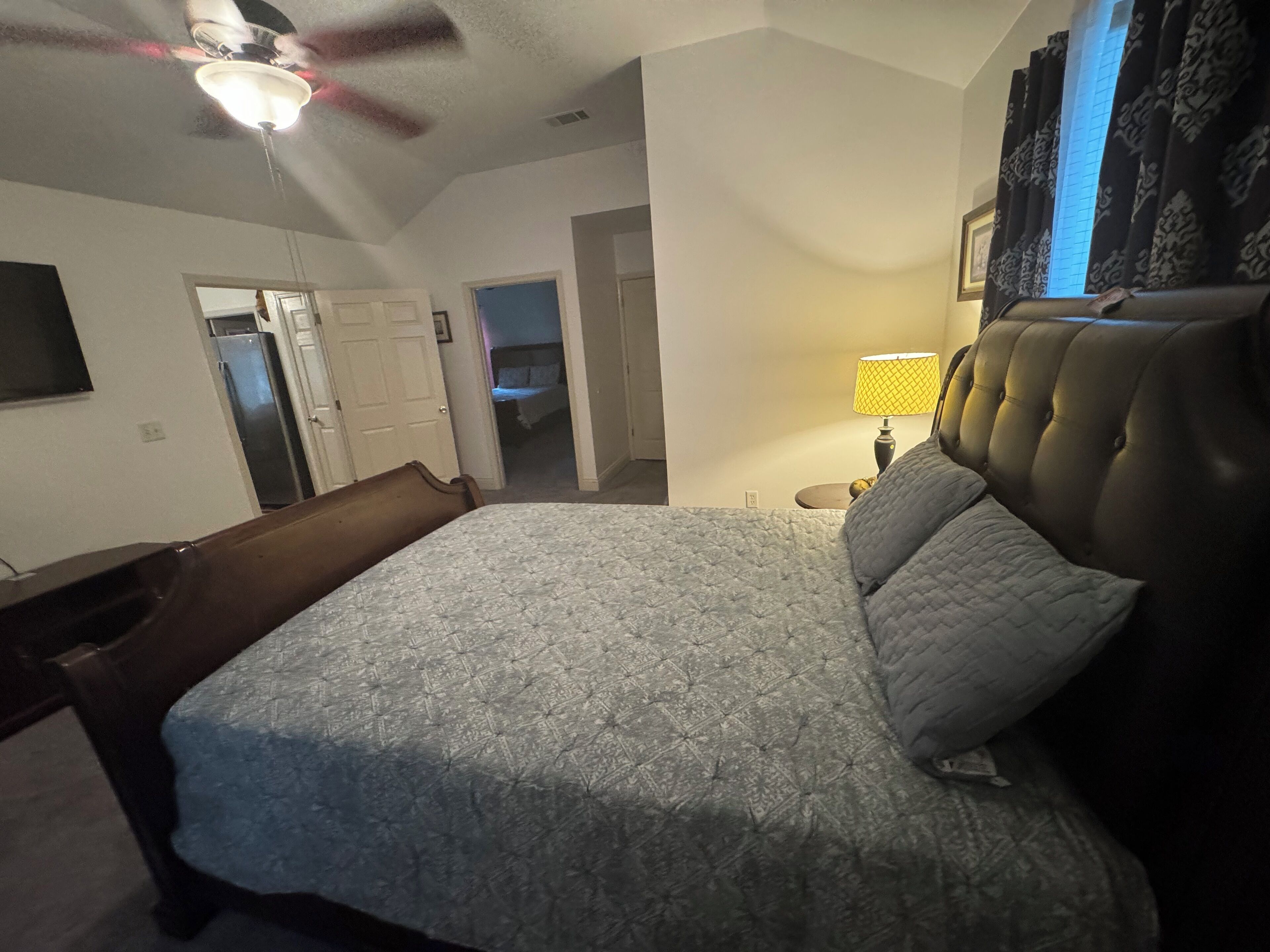 Un dormitorio con una cama, ventilador de techo, lámpara y vista a un pasillo.