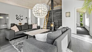 A modern living room with a sectional sofa, a round chandelier, a fireplace, and a view of greenery through a window.