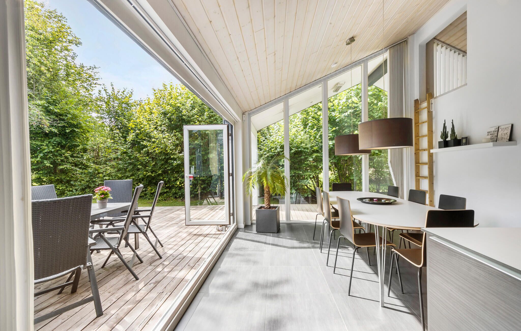 Ein offener Wohnbereich mit Holzterrasse, einem Essbereich mit langem Tisch und Stühlen sowie großen Fenstern mit Blick ins Grüne.